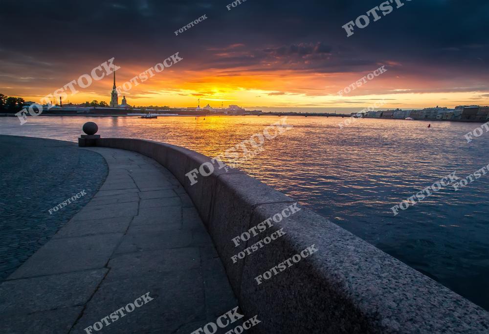 На фотографии запечатлен закат над Невой с видом на Петропавловскую крепость. Оранжевые и пурпурные оттенки неба отражаются на воде, создавая теплую и спокойную атмосферу. Каменная набережная изгибается вдоль реки, подчеркивая перспективу вида. Этот пейзаж передает романтику и величие вечернего Санкт-Петербурга.